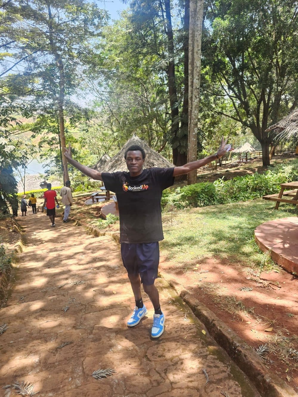 Jo Mogere wearing the black BookedUp Africa t-shirt with arms raised on a stone path through trees at a lodge with thatched huts